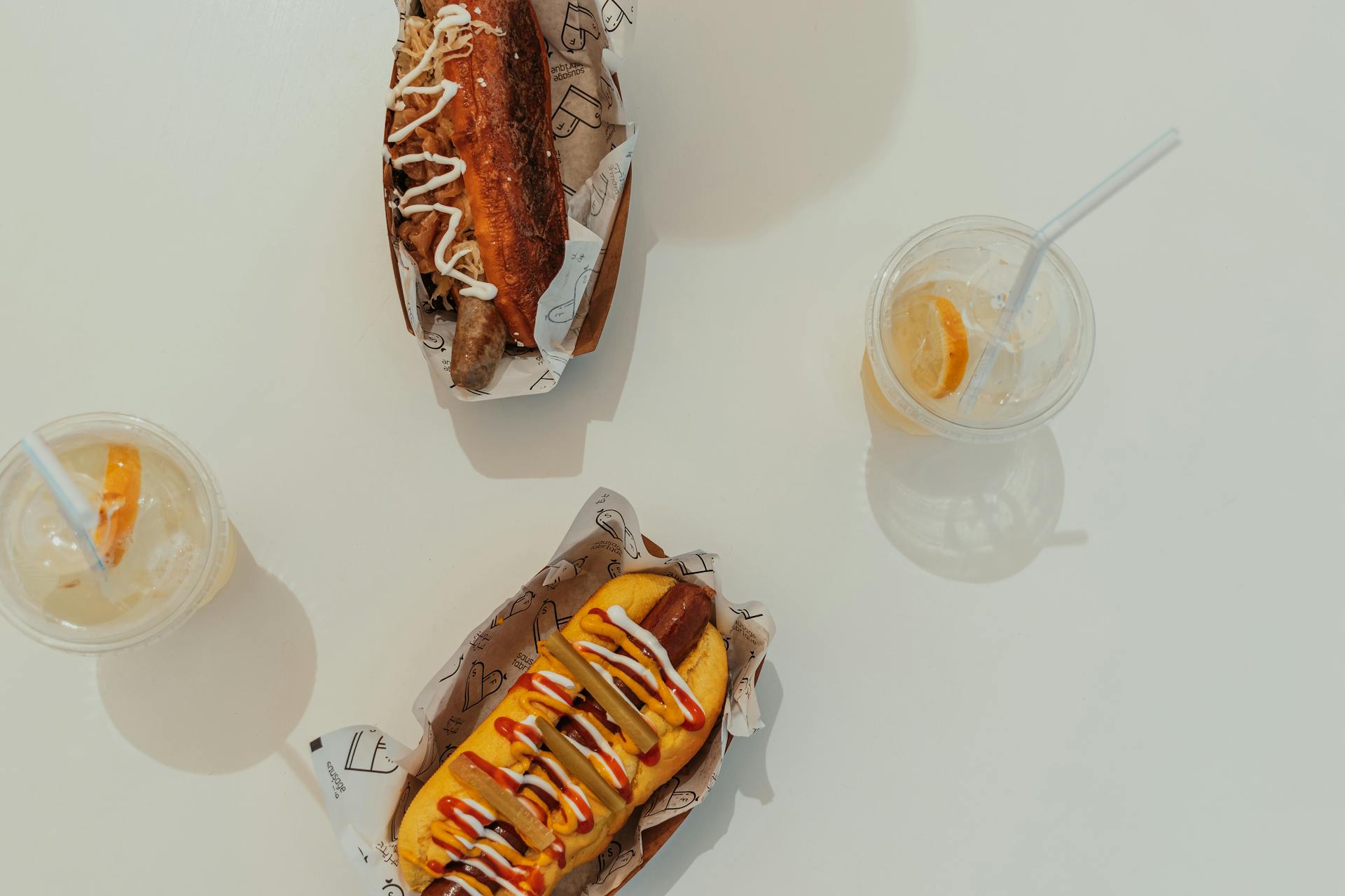 Two gourmet hot dogs with toppings and iced lemonade on a white table in a sunny outdoor setting