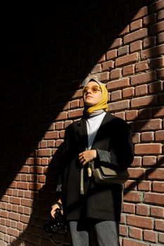 A woman in a yellow hijab and sunglasses stands with a camera in sunlight.