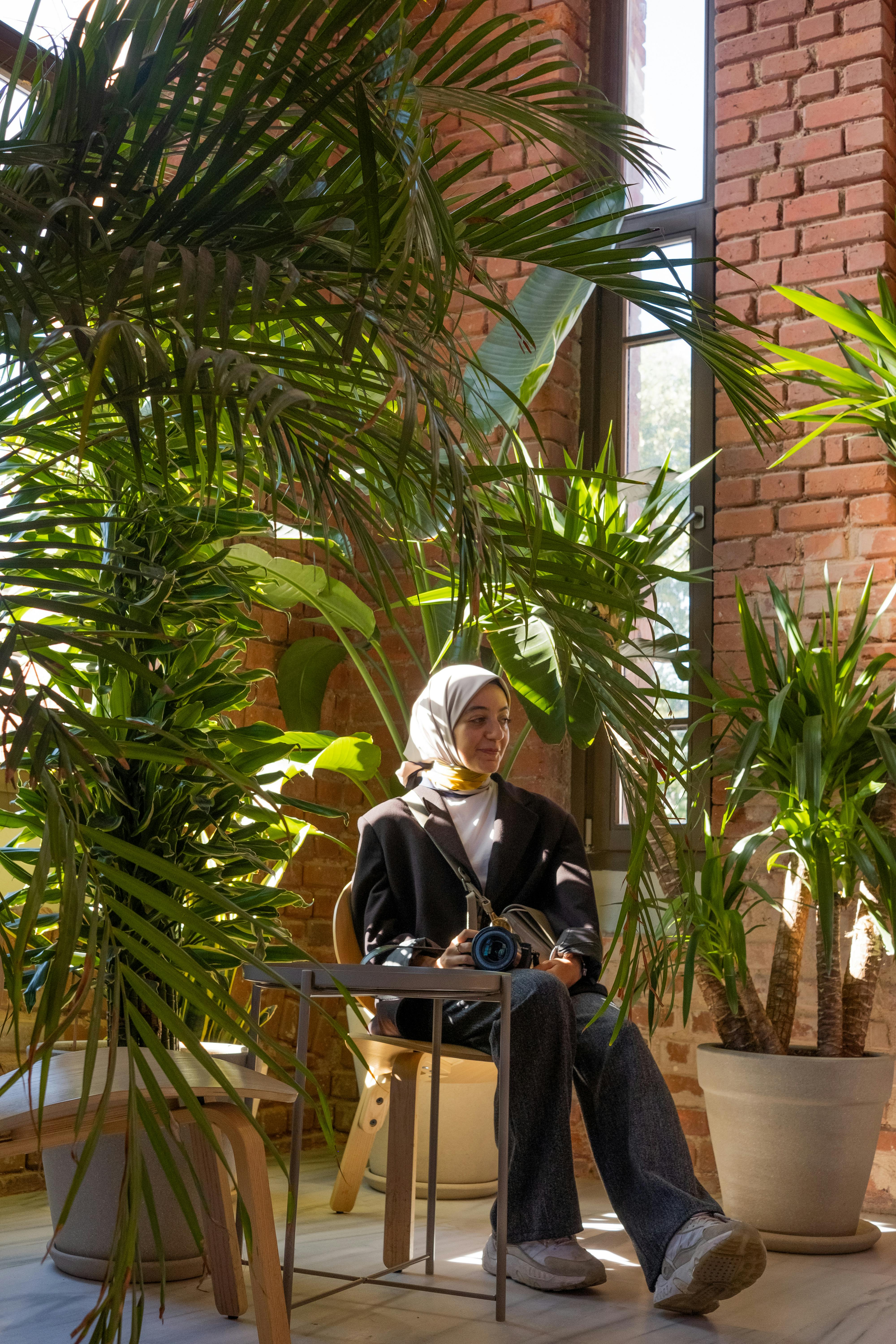 Young woman seated in a vibrant indoor garden surrounded by tropical plants and soft sunlight.