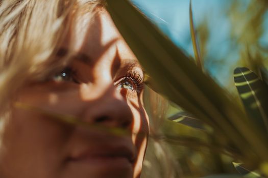 Woman's face with vibrant eyes surrounded by tropical leaves in sunlight.