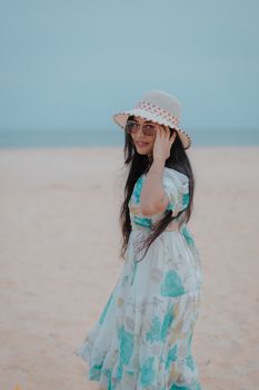 A fashionable young woman enjoys a sunny day at Kanniyakumari Beach, India, wearing a floral dress and sun hat.