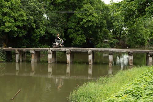 Free A person cycles over an old stone bridge in a lush green landscape with a calm river reflection. Stock Photo