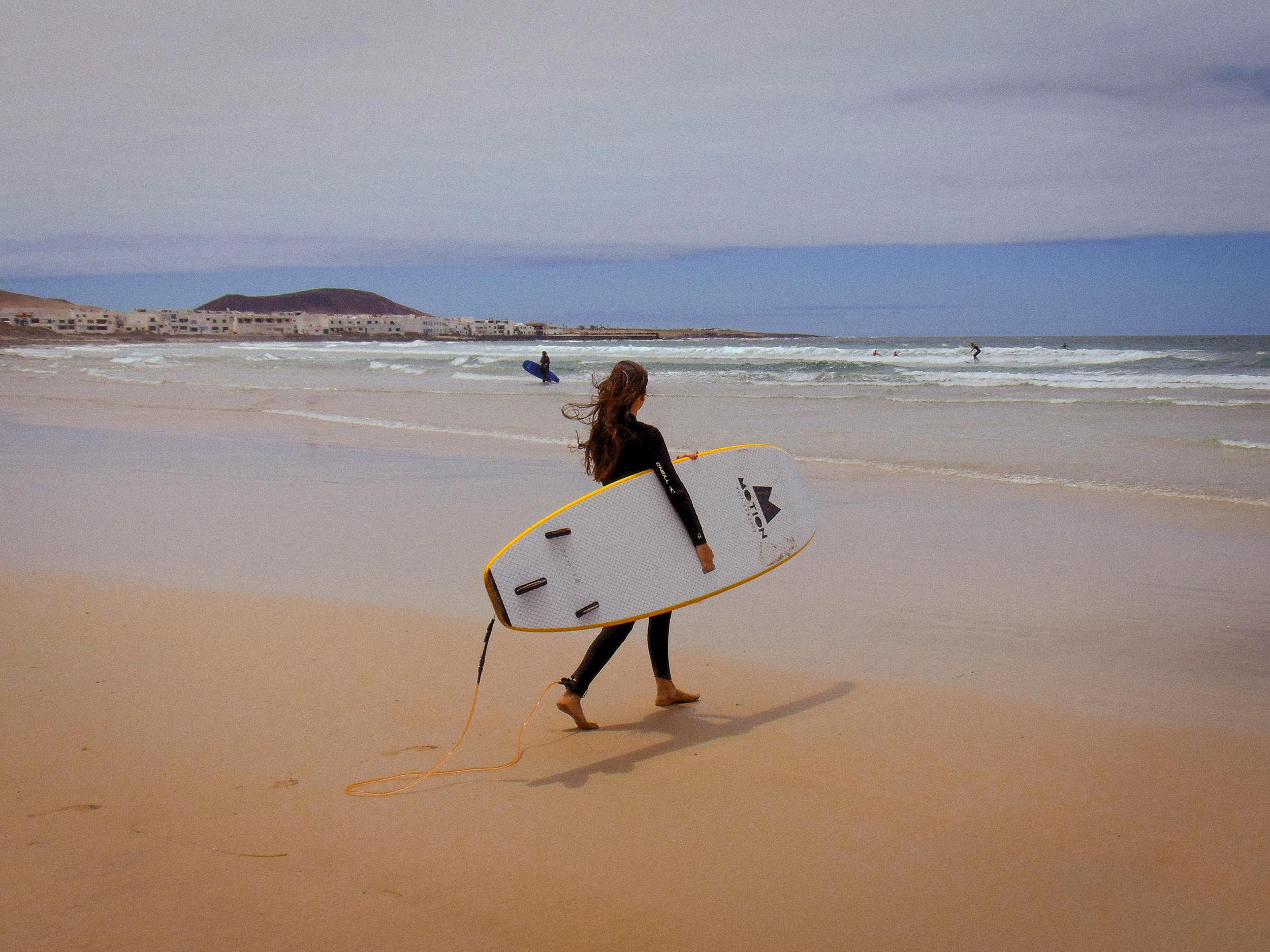 A lone surfer carrying a board along Caleta de Famara beach in Spain, perfect for summer recreation.