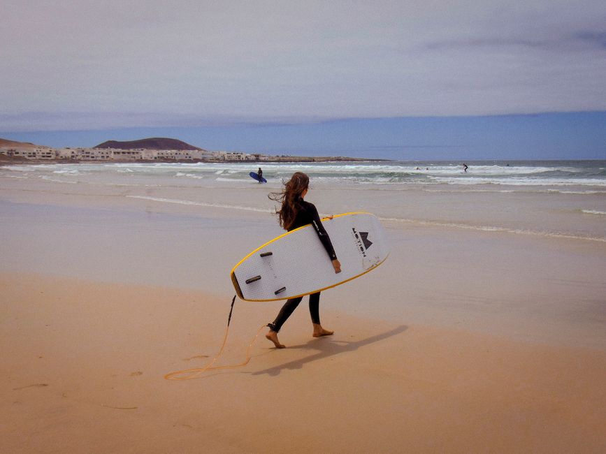 A lone surfer carrying a board along Caleta de Famara beach in Spain, perfect for summer recreation.