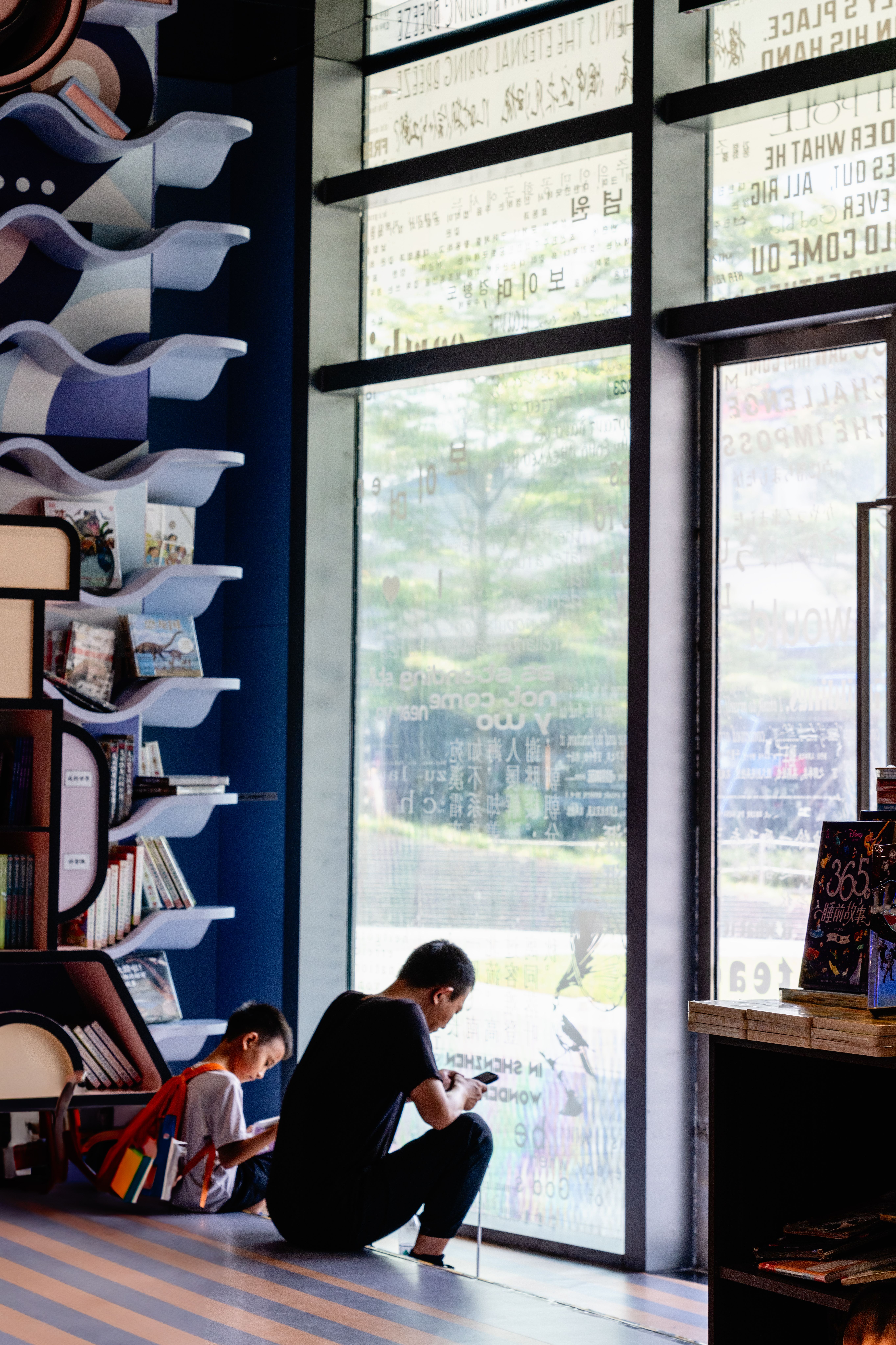 A man and child reading by a large window in a modern library with bookshelves.