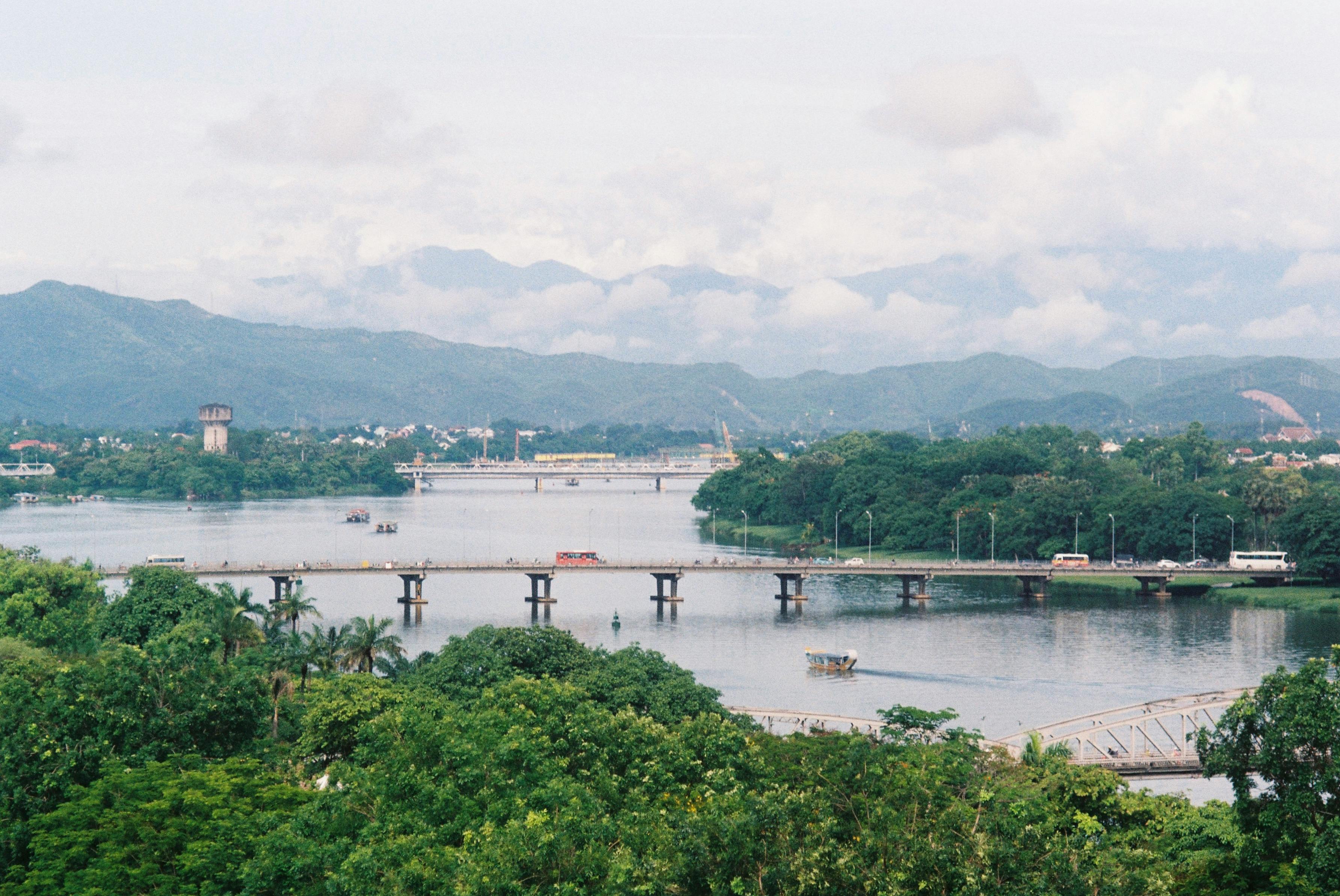 Picturesque view of a river with bridges, lush greenery, and distant mountains.