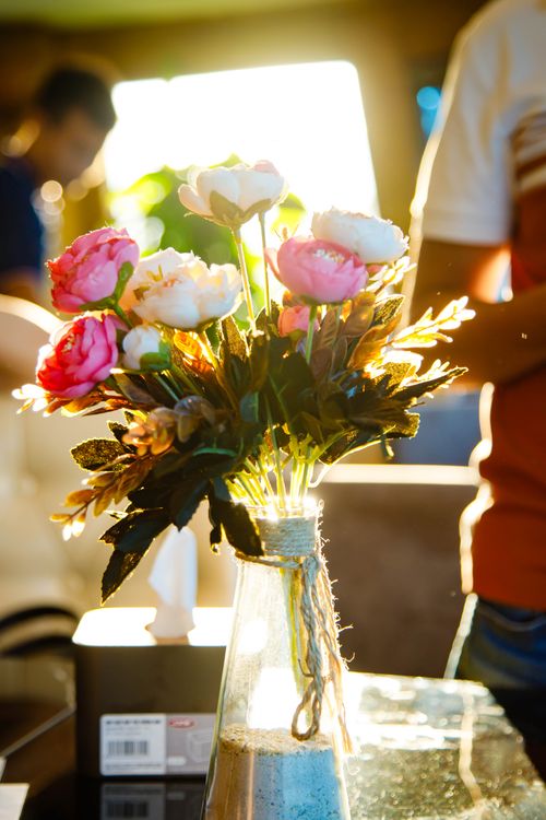 Free A bouquet of artificial roses in sunlight on a glass table, with people blurred in the background. Stock Photo