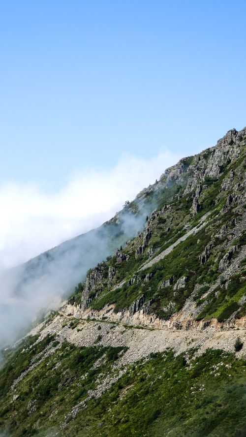 Free Breathtaking view of a mountainside pathway enveloped by mist and clouds under a clear sky. Stock Photo