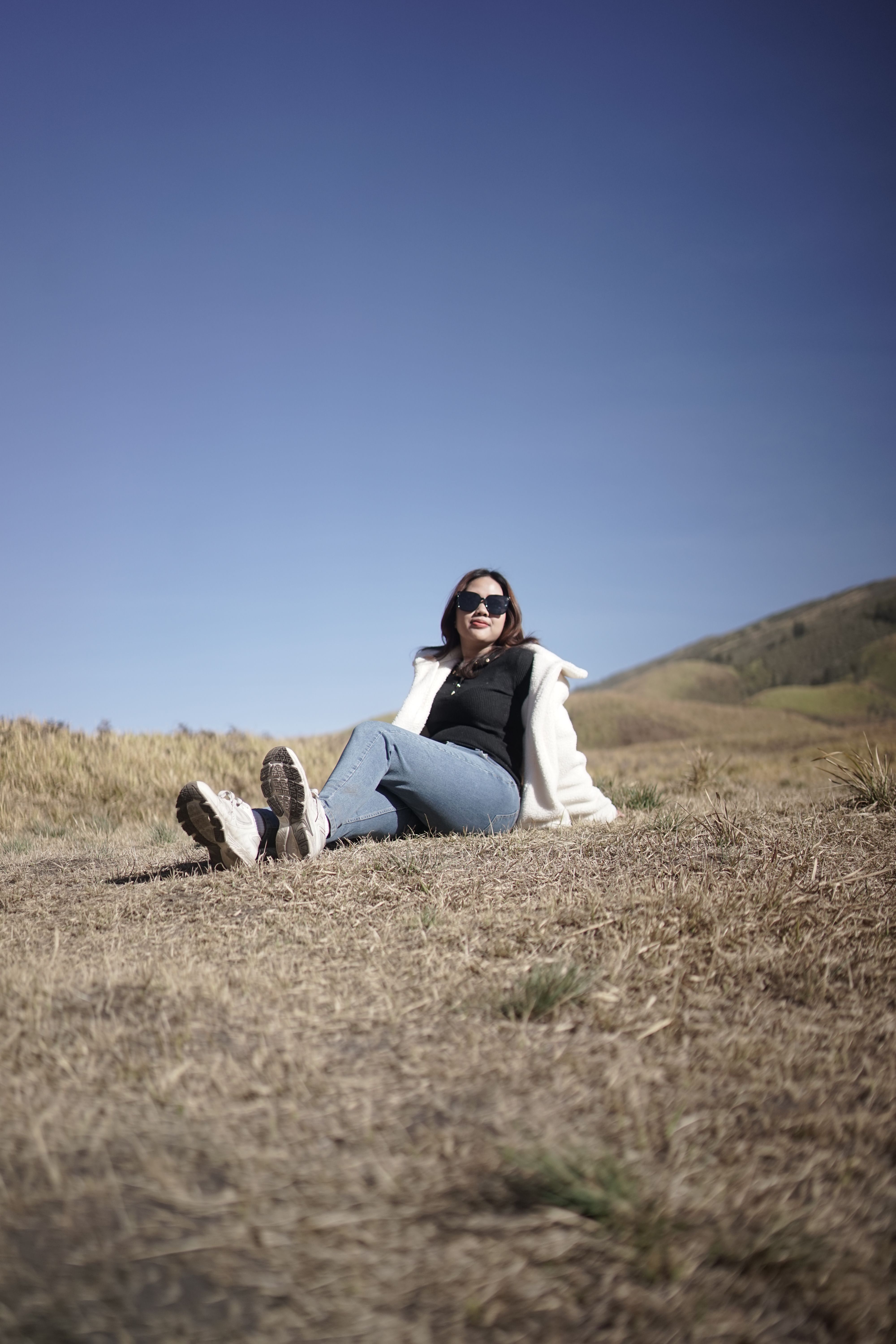 A young woman enjoys a peaceful day sitting on a grassy hillside under a clear blue sky.