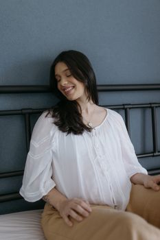 Content woman in casual clothing smiles while sitting on the bed in a cozy bedroom.
