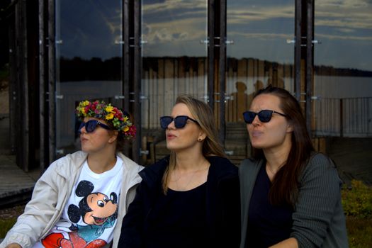 Three women wearing sunglasses pose in front of a modern glass building, enjoying an outdoor urban scene.