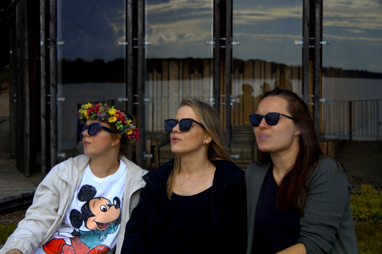 three women wearing sunglasses pose in front of a modern glass buildin