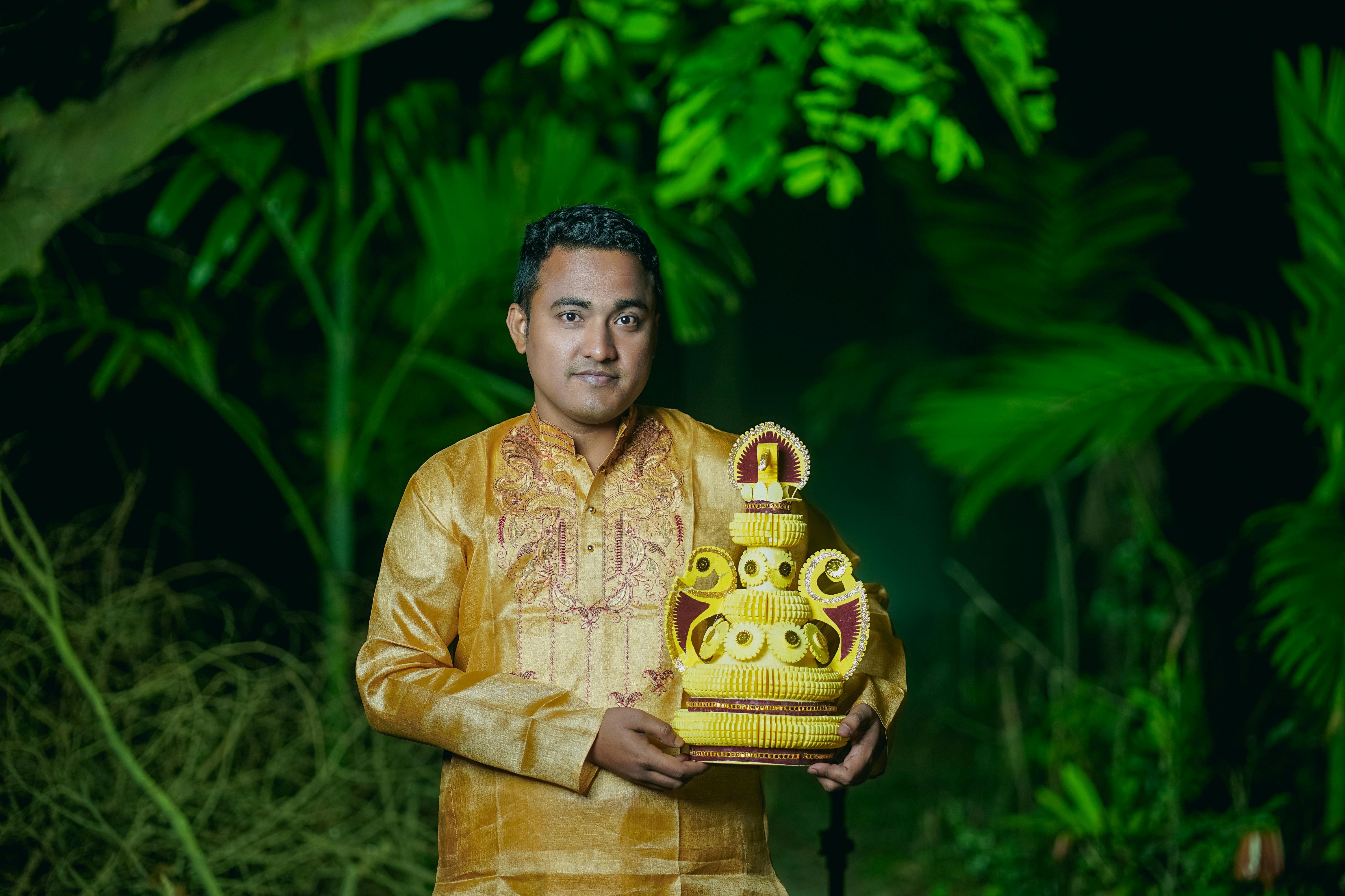 Man in traditional Bangladeshi attire holding decorative art outdoors in lush green setting.