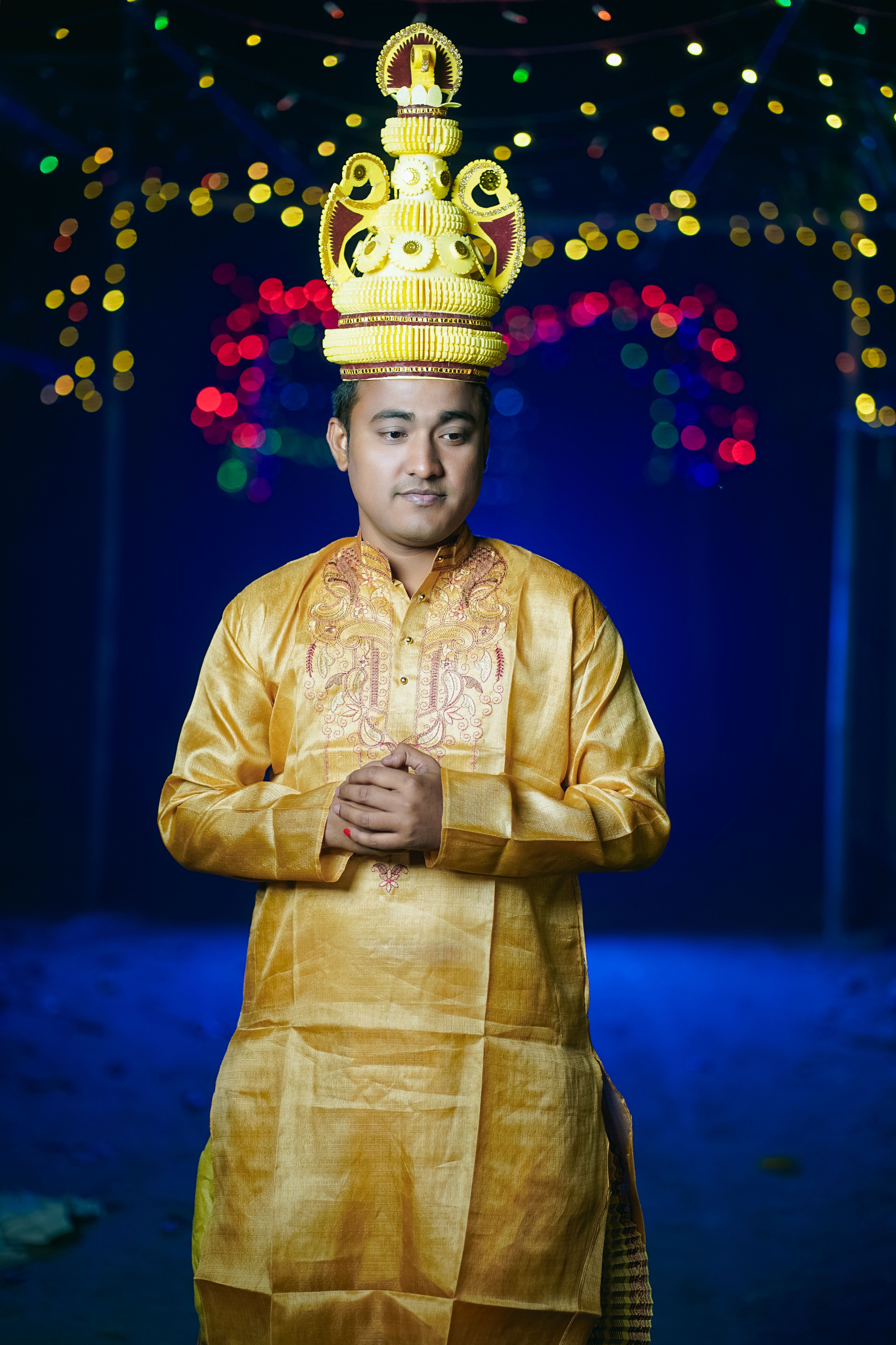 A man in traditional Bengali wedding attire with ornate hat under colorful lights.