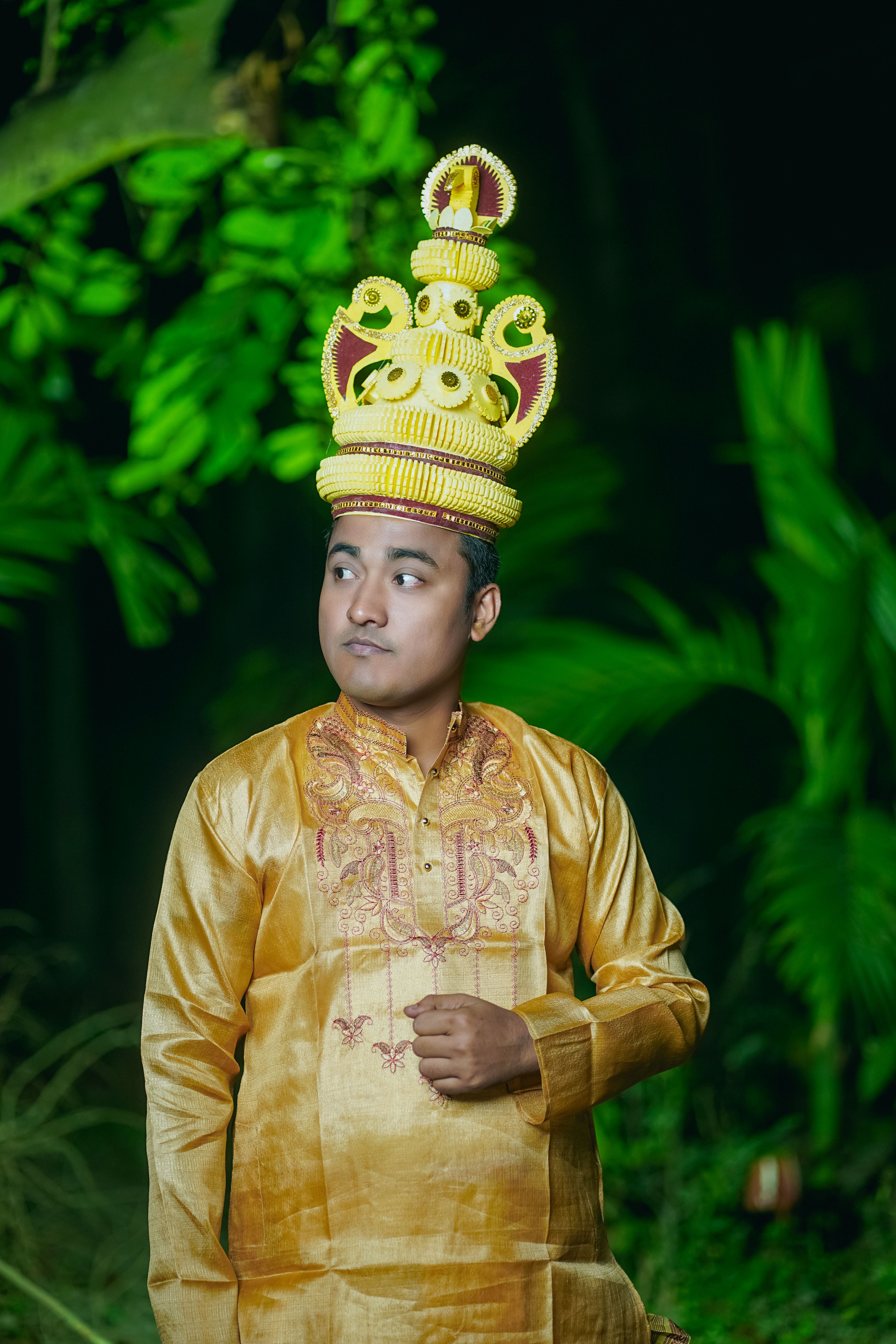 A man in elegant traditional Bangladeshi attire and a crown, posed outdoors against lush greenery.
