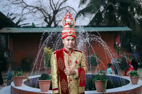 Free A groom dressed in traditional Bangladeshi attire stands proudly outdoors with a fountain backdrop. Stock Photo