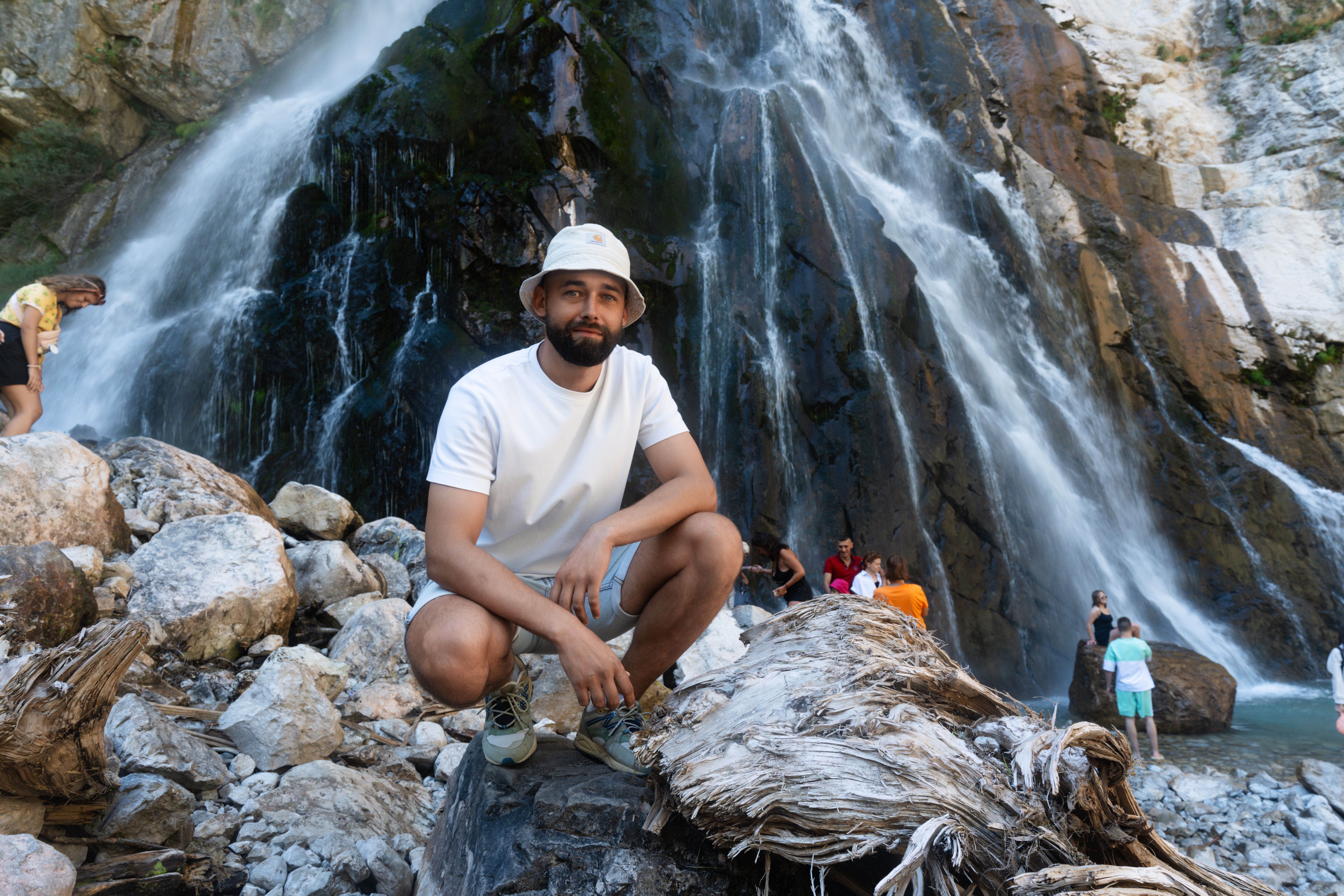 A young man in casual attire poses near a stunning waterfall in an outdoor adventure setting.
