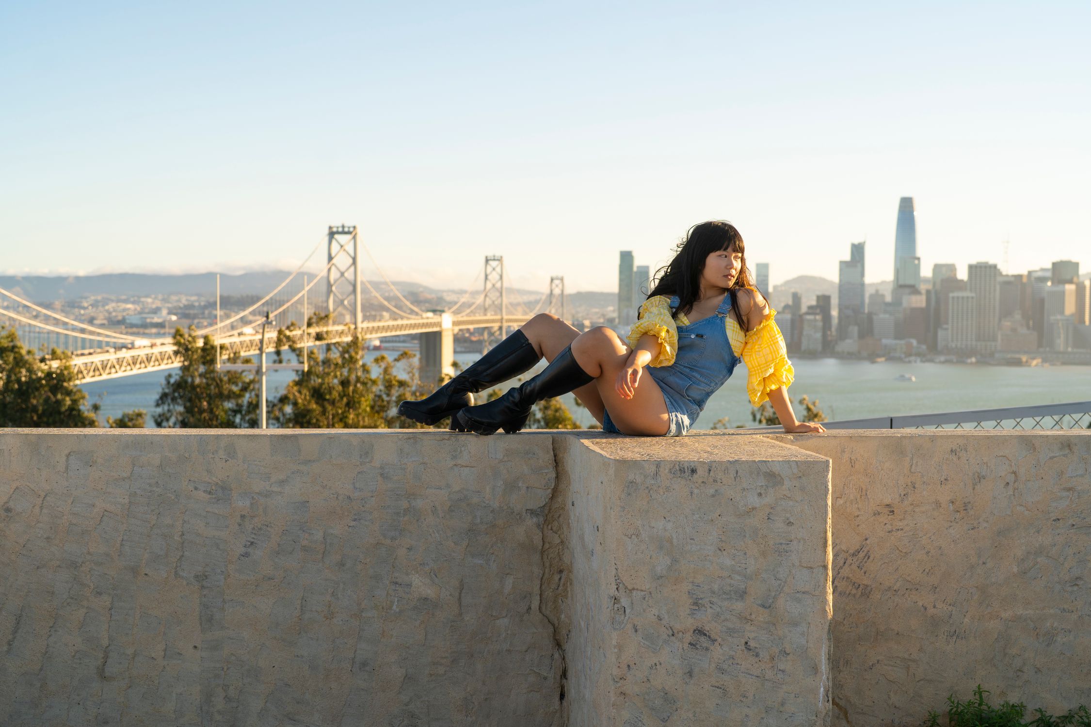 Young woman sitting on a ledge overlooking San Francisco and the Bay Bridge at sunset.