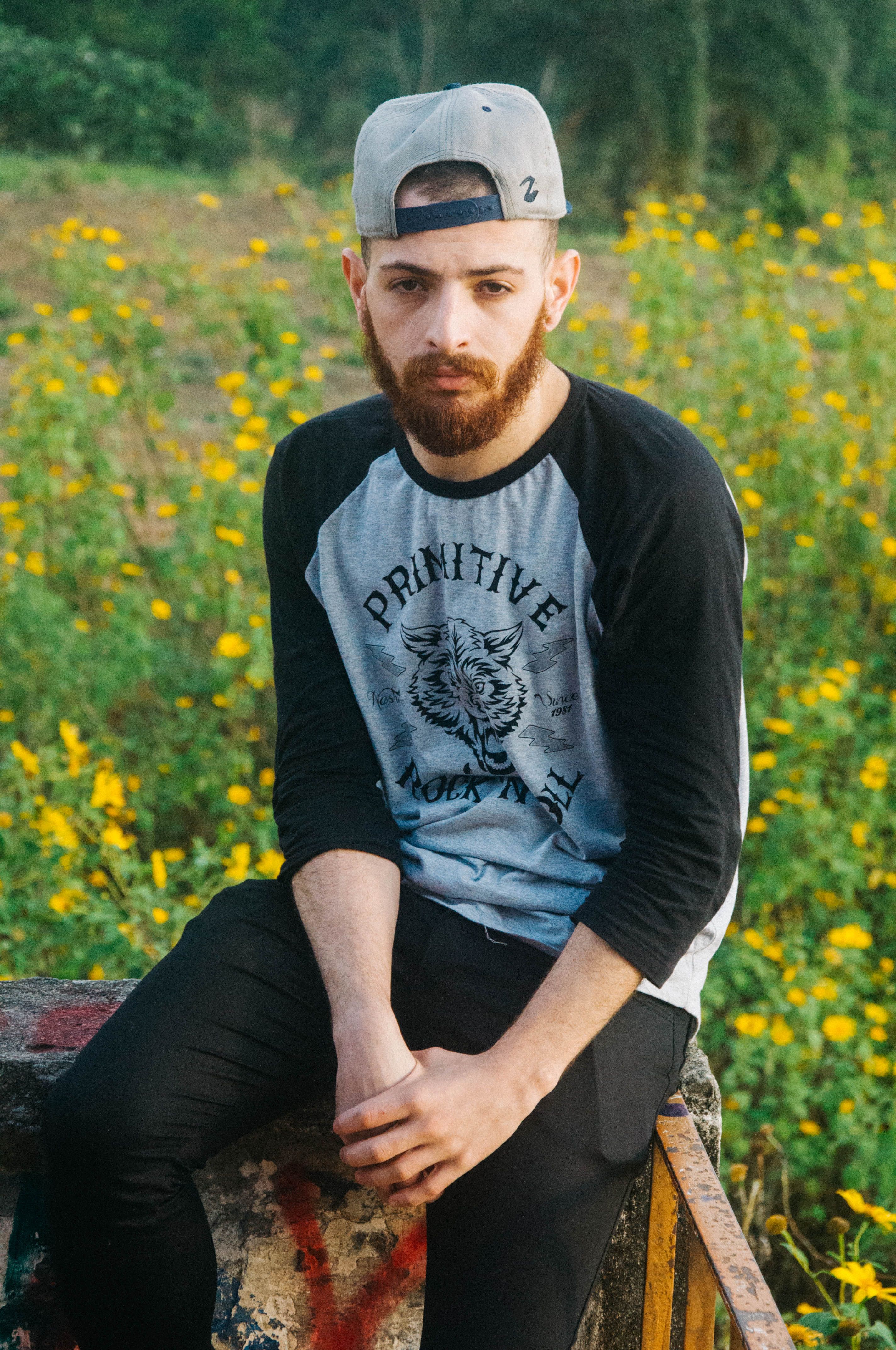 Young man in casual wear poses in a sunny, flower-filled countryside setting.