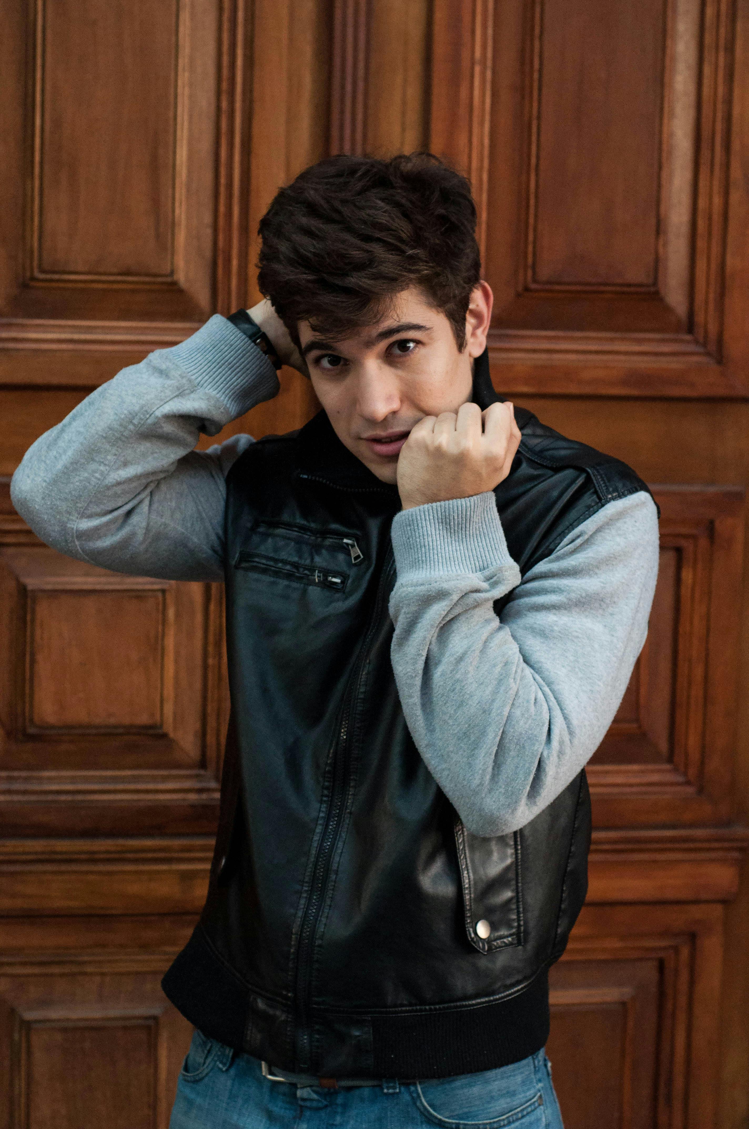 Portrait of a young man adjusting his leather jacket indoors, against a wooden background.