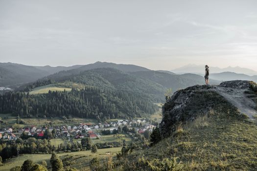 A hiker stands on a cliff overlooking a serene valley and small town during daylight.