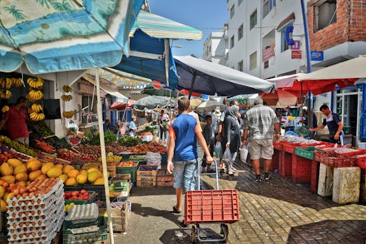 Bustling outdoor market scene with people shopping for fresh produce under colorful umbrellas.