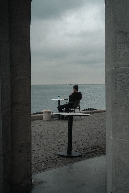 Free Man sitting alone at a seaside cafe table, gazing at the ocean under a cloudy sky. Stock Photo