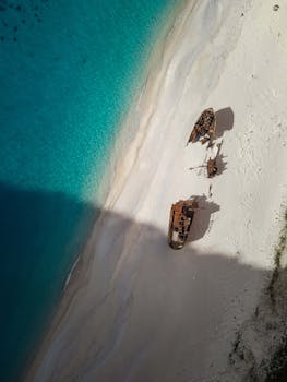 Aerial shot capturing rusted shipwrecks on a white sandy beach beside vibrant blue waters.