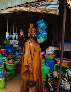 A woman in traditional African attire stands amidst colorful market goods, showcasing vibrant culture.