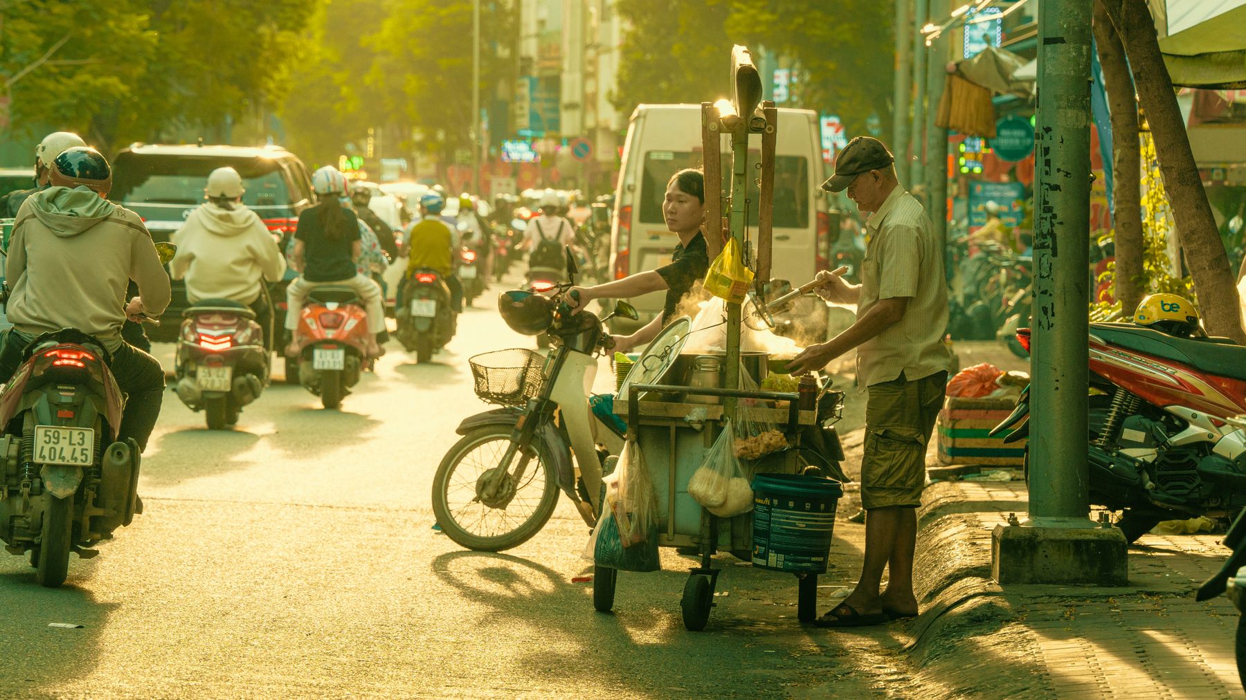 Crowded city street with people on motorcycles and a street food vendor serving customers under warm sunlight.