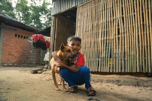 Free A cheerful child joyfully embraces a dog outdoors, surrounded by rustic bamboo architecture. Stock Photo