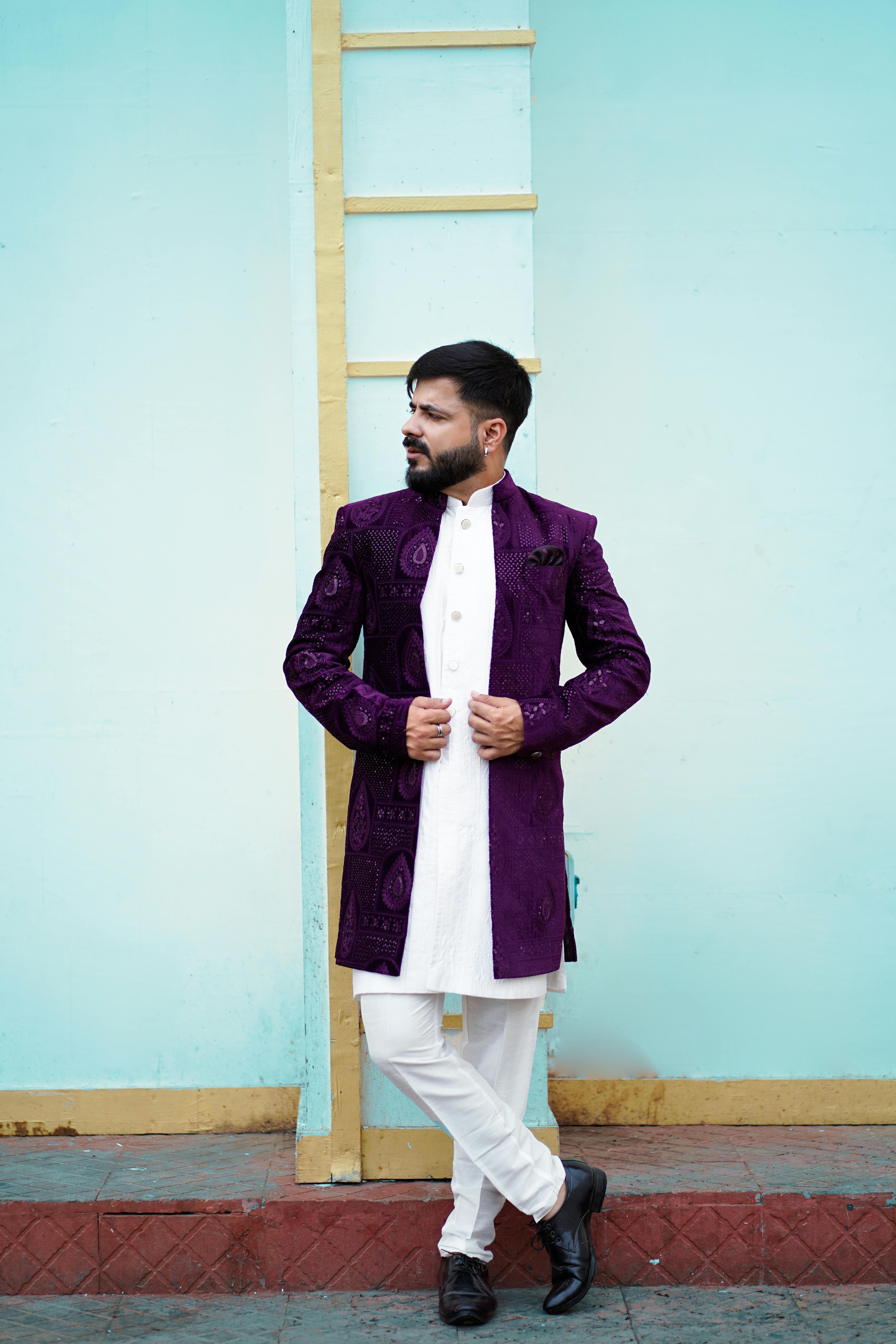 Man in vibrant Indian attire posing confidently outdoors with a light blue wall backdrop.