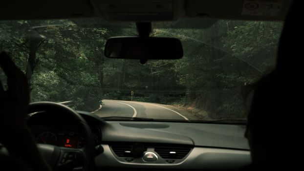Interior view of a vehicle driving along a winding forest road, capturing a rainy, moody atmosphere.