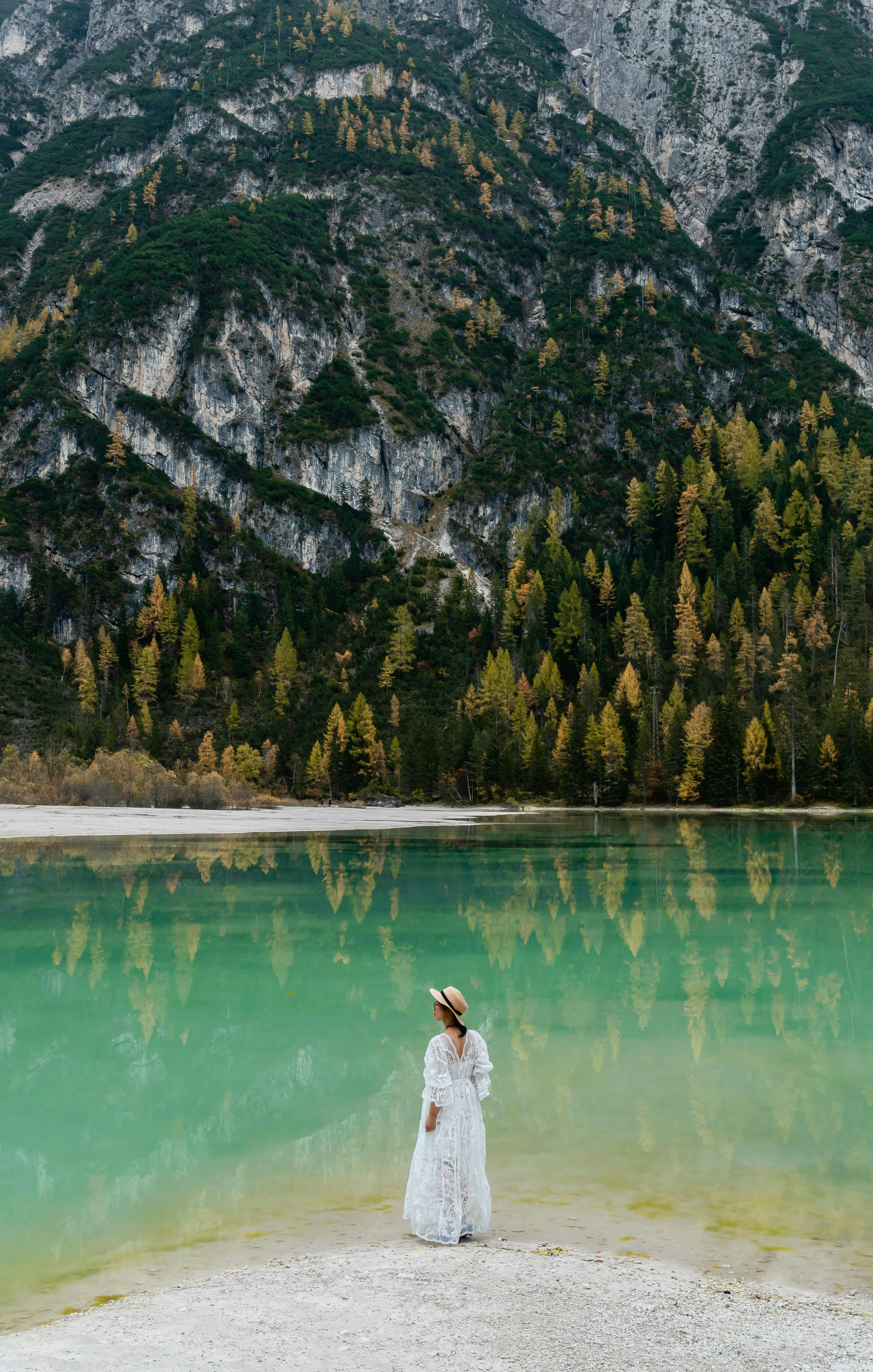 A woman stands by a tranquil mountain lake with forest and cliff reflections, creating a serene autumn landscape.