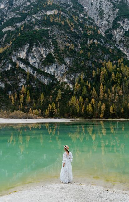 A woman stands by a tranquil mountain lake with forest and cliff reflections, creating a serene autumn landscape.