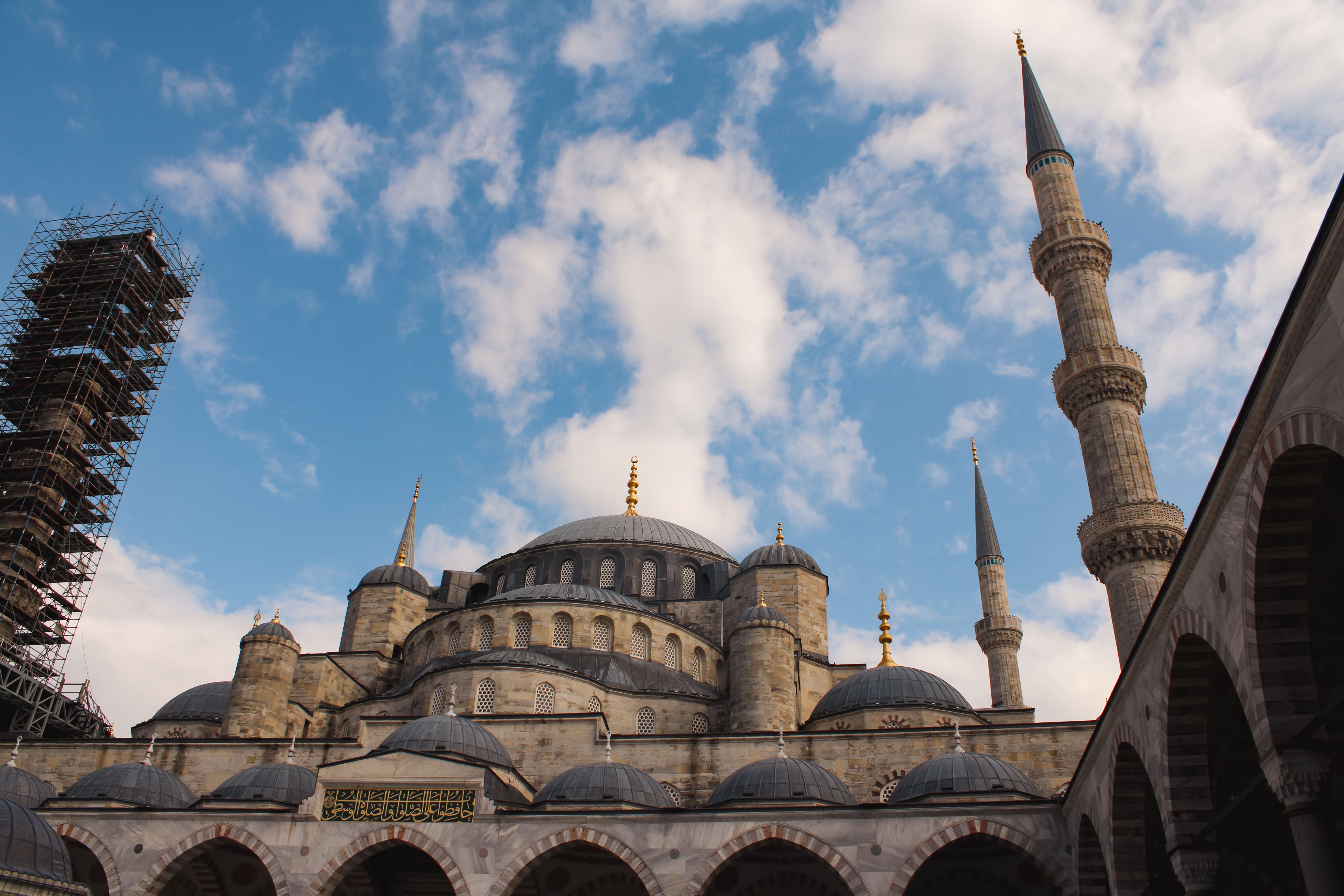 A large grey-domed mosque with half-domes and tall minarets, seen over the arches of a courtyard with scaffolding at the left edge