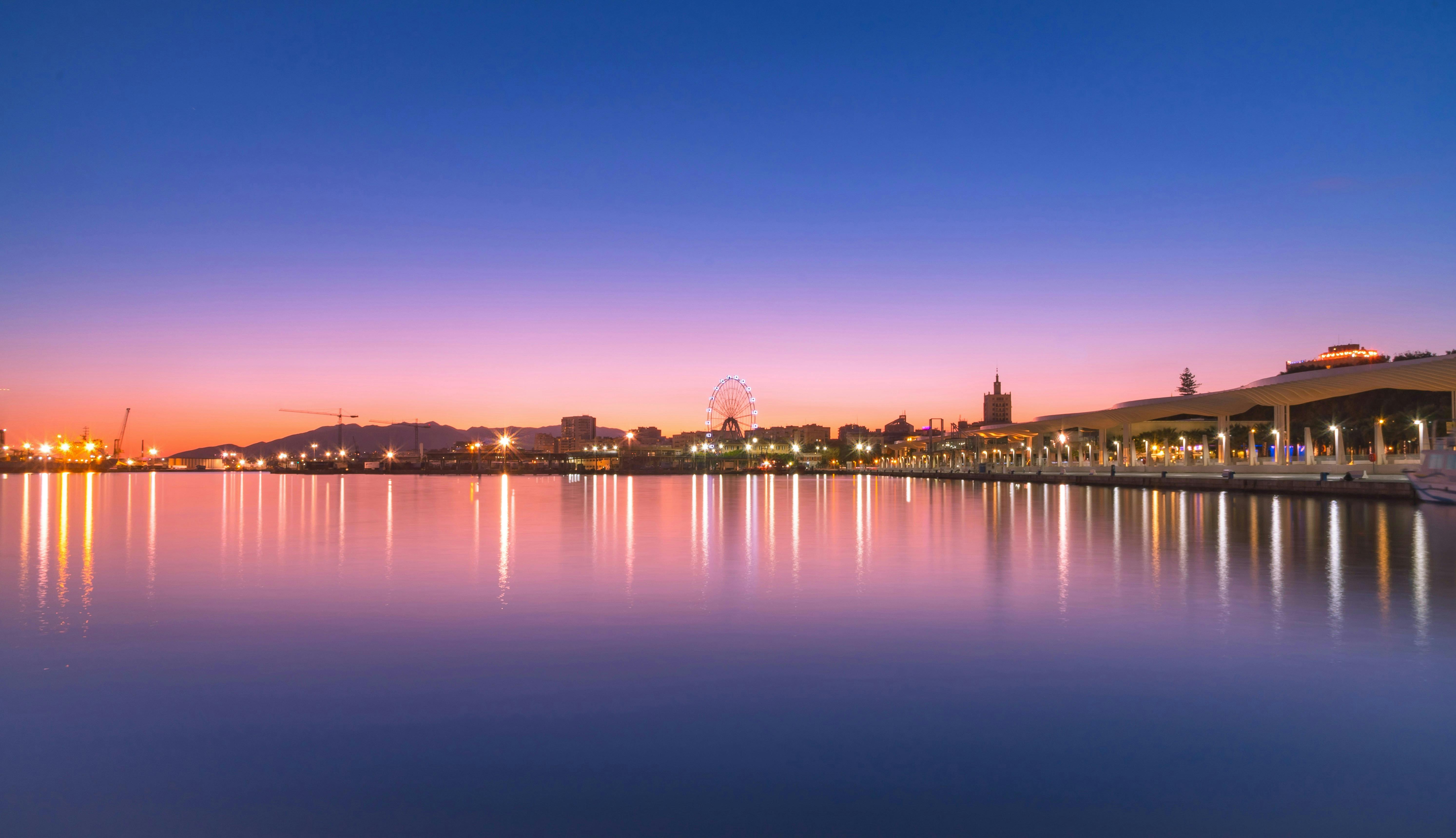 Scenic city skyline reflecting in calm water at sunset, showcasing a Ferris wheel and vibrant colors.