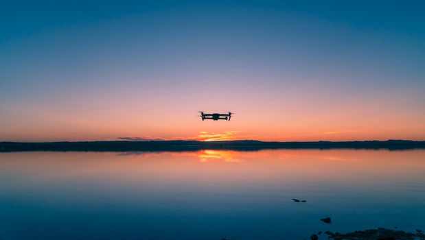 Stunning sunset silhouette of a drone hovering over a calm lake reflection.