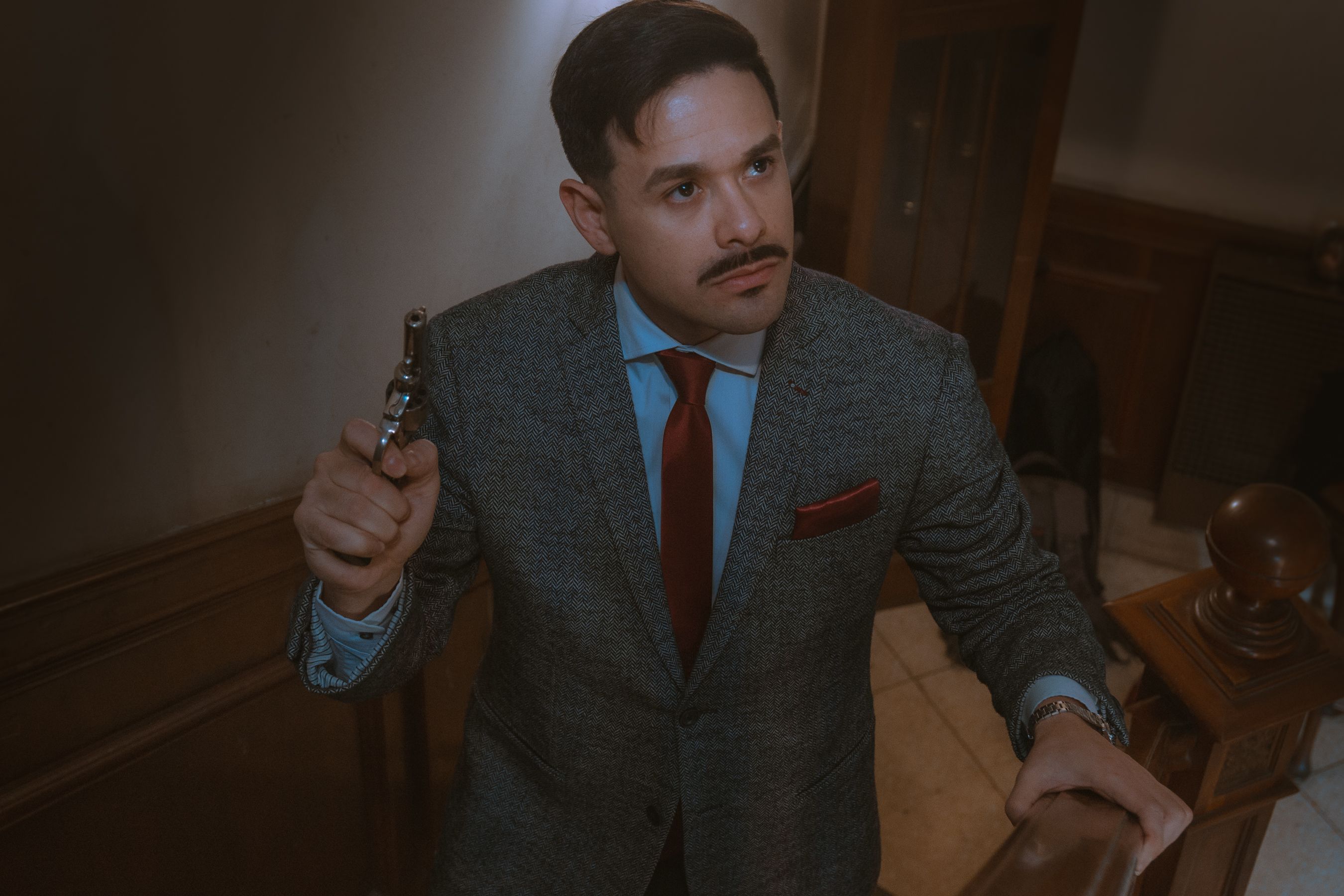 A fashionable man in a suit and tie holding a revolver, standing in an indoor setting.