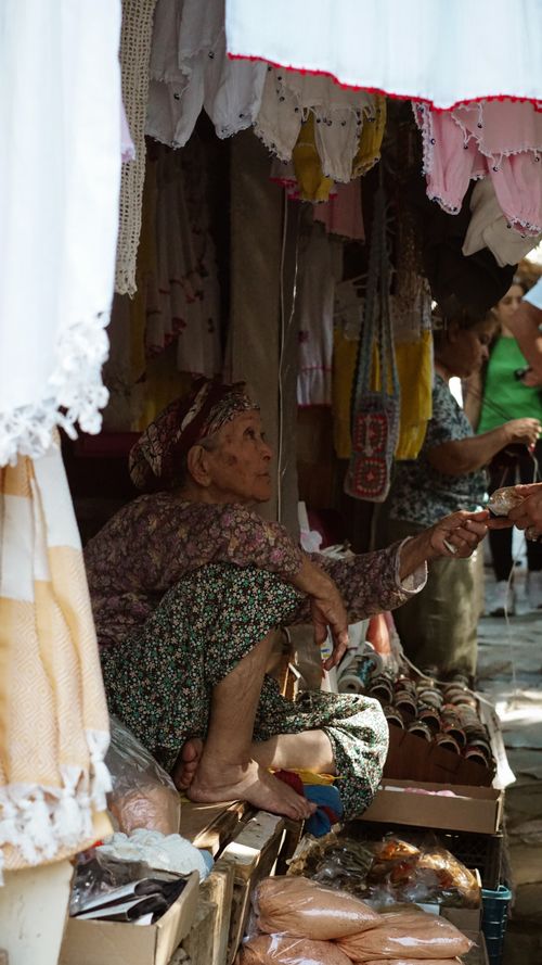 Free An elderly woman in traditional clothing sells handmade goods at an outdoor market stall. Stock Photo