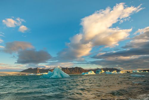 Free Scenic view of icebergs floating in a vast ocean with mountains and a dramatic sky at sunset. Stock Photo