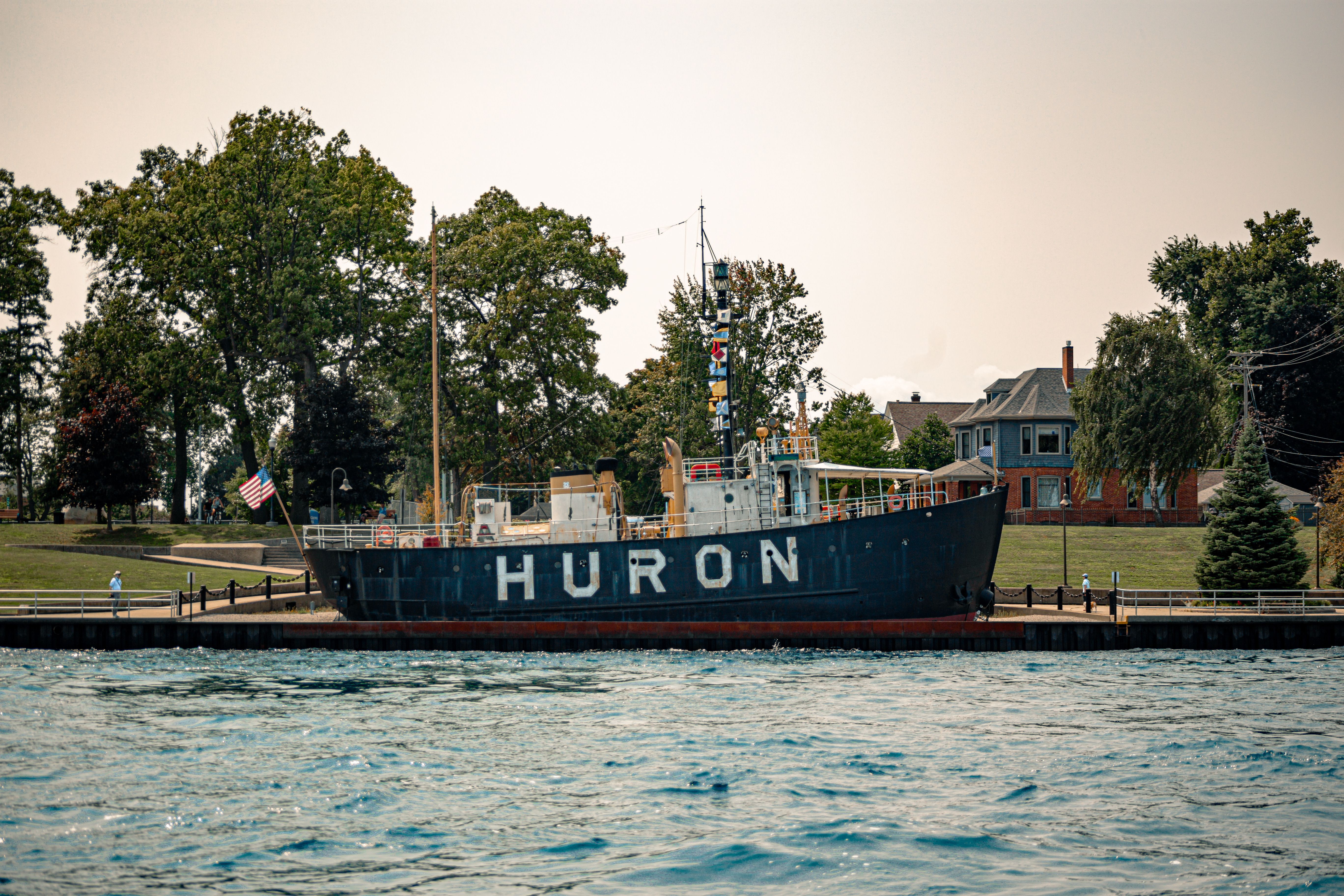 Huron lightship docked on a serene waterfront with trees and historic buildings in the background.