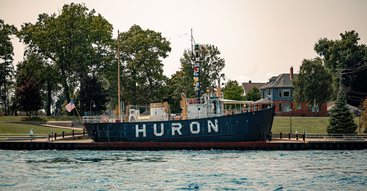 Huron lightship docked on a serene waterfront with trees and historic buildings in the background.
