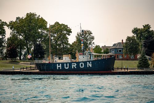 Free Huron lightship docked on a serene waterfront with trees and historic buildings in the background. Stock Photo