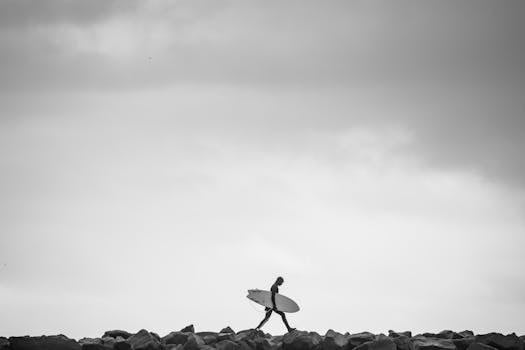 Black and white image of a surfer walking with a surfboard over rocks against a cloudy sky.