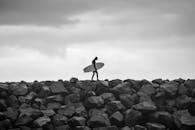 A black and white photo of a surfer walking on rocks