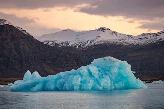 A stunning icy landscape with a blue iceberg against snow-capped mountains at sunset.