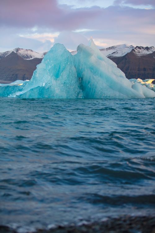 Free Majestic iceberg floating in serene Arctic waters with snow-capped mountains at sunset. Stock Photo