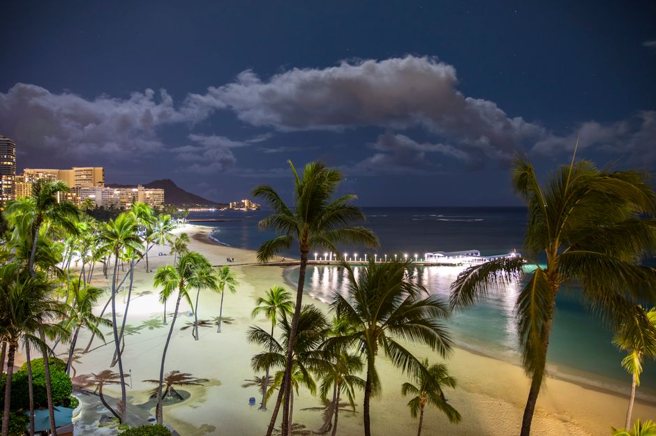 Stunning night view of Waikiki Beach with palm trees, pier, and city lights under a starry sky.
