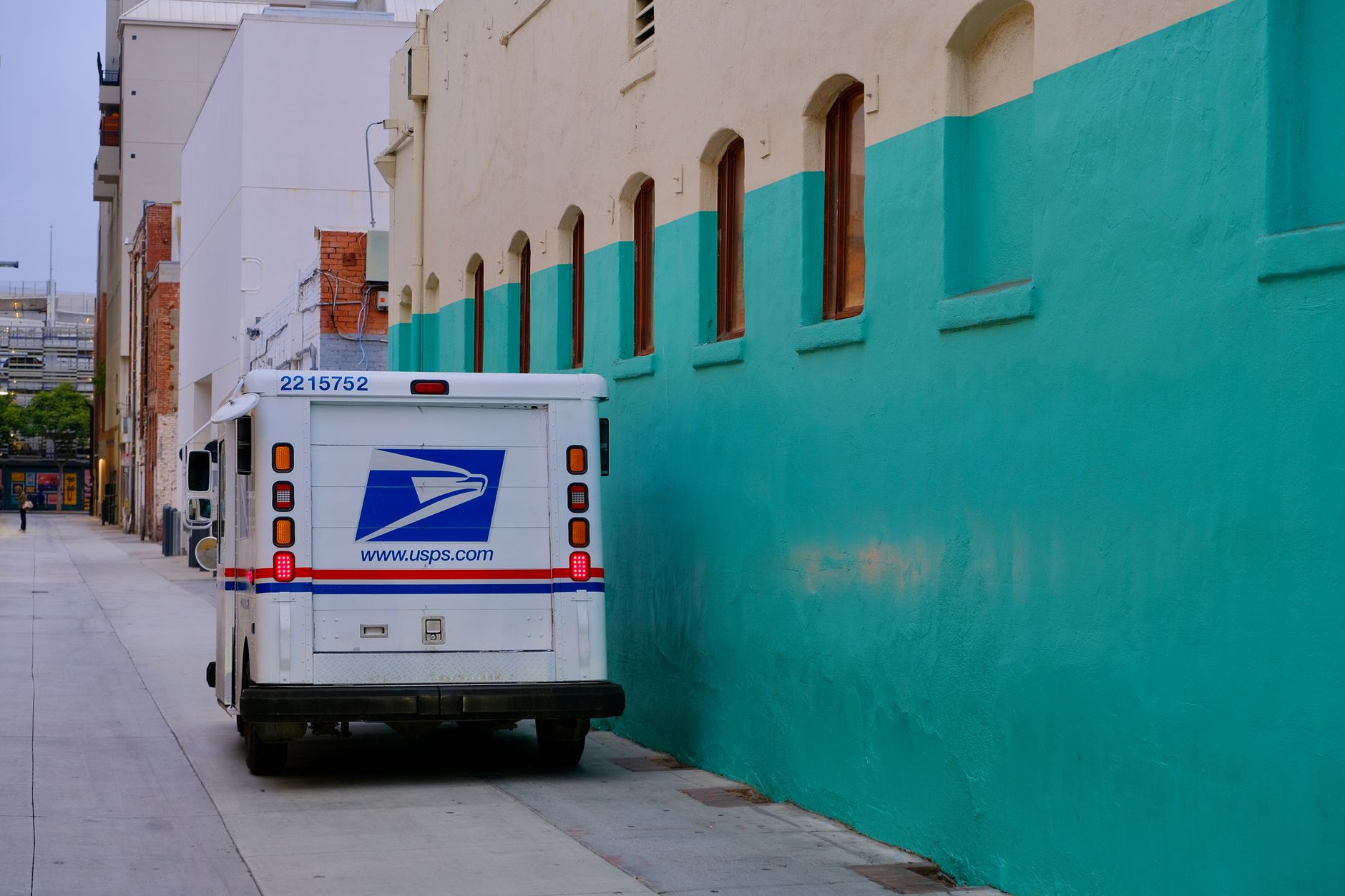 A USPS delivery truck parked in a vibrant urban alley beside colorful architecture.